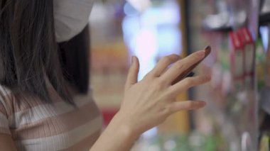 Tıbbi maskeli Asyalı bir kadın aradığı malzemeleri bulmak için akıllı telefon kullanıyor, süpermarkette rafların yanında duruyor, internette sörf yapıyor, telefonları kaydırıyor, virüs salgınını önlüyor.