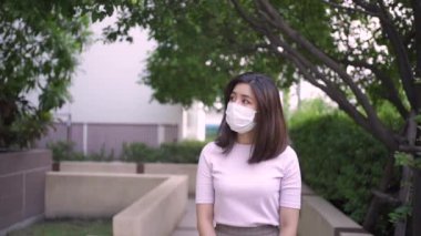 Genç, Asyalı bir kadın beyaz maske takıyor. Açık hava parkında, ağaç gölgelerinin altında kameraya doğru yürüyor. Yalnız ev karantinası, kovid19 Corona virüsü.
