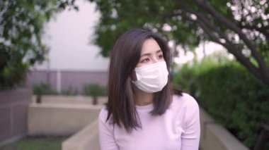 Genç Asyalı kadın maske takıyor. Yavaş çekimde kameraya doğru yürüyor. Açık hava parkında ağaçların altında yürüyor, kendini karantina altında, kovid19 Corona virüsü.