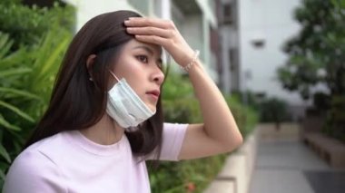4K Asyalı genç kadın oturuyor ve koruyucu maske takıyor, kirlilik virüsü problemi, modelin arkasındaki yeşil yapraklar ve çalılar, covid19 Corona virüsü, mahallesinde güneşli bir gün, sağlık konsepti