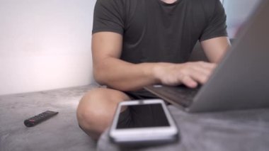 Asyalı erkek, akıllı telefonlu dizüstü bilgisayar kullanıyor, internet üzerinden çalışma programı yapmak için kişisel kablosuz bağlantı noktası kullanıyor, klavyeye oturuyor, kanepede oturuyor, yarışları başarıyla kazanıyor. 