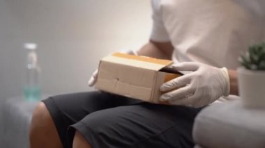 İnsan müşteri küçük karton kutuları açıyor, evdeki koltukta boks yapıyor, Asyalı erkek tüketici küçük karton paketleri açıyor, şaşırtıcı bir şekilde iyi satın alınmış posta teslimatı, covid-19 krizi, ekspres sevkiyatı alıyor.