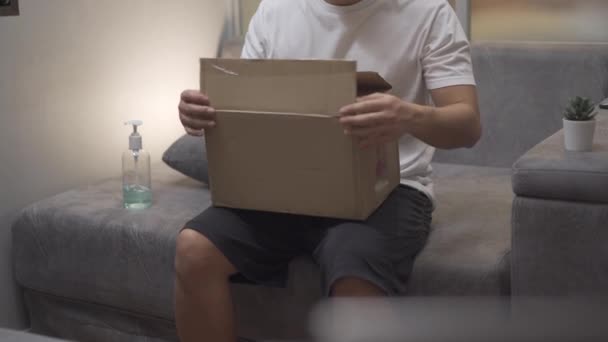 亚洲男性无盒装纸板箱 在家中灰色沙发上 邮件接收机打开小纸箱包裹 包裹19危机 包裹递送网上购物 航运邮件业 — 图库视频影像
