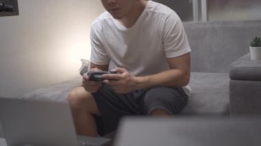 Asyalı adam konsol oyunu oynuyor. Oturma odasındaki gri koltukta oturuyor. Neşe kontrolörüyle oyun oynuyor, yaşam tarzını karantinaya alıyor.