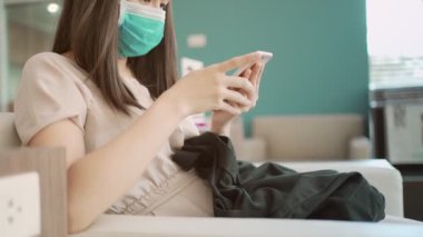 4K Asyalı kadın akıllı telefon kullanarak maske takıyor, kadın hastane koridoru lobisinde bekliyor, klinik tesisinde online sağlık makaleleri okuyor, kendini hastalıktan koruyor, hayat sigortası reklamı yapıyor.