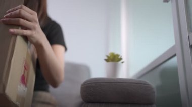 Asyalı bir kadın karton kutuları açıyor, koltukta oturuyor, alıcısı küçük karton paketleri açıyor, covid-19 krizi, paket teslimatı çevrimiçi alışveriş, paketlerin açılması evdeki kanepede oturuyor.