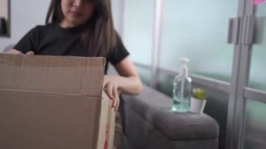 Mutlu Asyalı kadın açık karton kutu paketi oturma odasındaki gri koltukta oturuyor, posta alıcısı karton paketi açıyor, covid-19 krizi, paket teslimatı çevrimiçi alışveriş, siyah gömlek