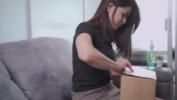 若いアジアの女性は自宅でソファの上に小さな段ボール箱のページの配信を開くと 受信機のアンパック小さなカートンの小包コンテナ Covid 19パンデミック 商品供給オンラインショッピング 電子商取引 — ストック動画