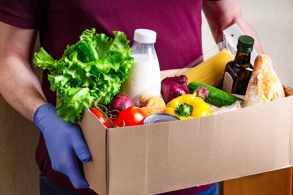 Volunteer in gloves holding food in a donation cardboard box with various food. Open cardboard box with oil, vegetables, milk, canned food, cereals and pasta. Food delivery concept with space for text