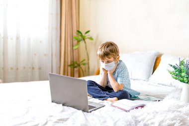 Uzaktan öğrenme çevrimiçi eğitim. Tıbbi maskeli hasta çocuk evde dijital tablet dizüstü bilgisayarla ders çalışıyor ve okul ödevi yapıyor. Eğitim kitaplarıyla yatakta oturmak.
