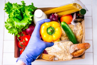 Eldivenli gönüllü bağış karton kutusunda çeşitli yiyeceklerle yemek veriyor. İçinde yağ, sebze, süt, konserve yiyecek, mısır gevreği ve makarna olan karton bir kutu açın. Yiyecek teslimatı kavramı .