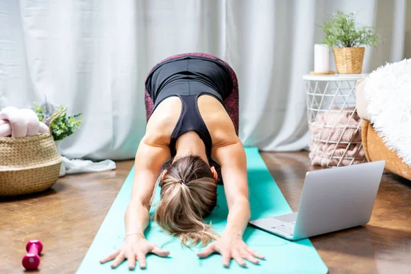 Young sporty attractive woman working out practicing yoga and using laptop and dumbbells at home in living room, doing Child exercise, Balasana. Концепция спорта и отдыха — стоковое фото