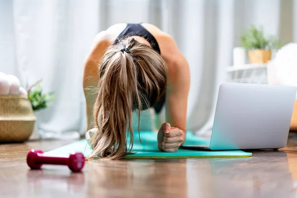 Молодая фитнес-женщина работает и с помощью ноутбука и гантели дома в гостиной, делая йогу или пилатес упражнения на бирюзовом коврике, стоя в позе доски. Концепция спорта и отдыха — стоковое фото