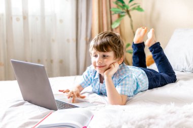 Uzaktan öğrenme çevrimiçi eğitim. Kafkasyalı mutlu çocuk elinde kitapla evde dijital tablet dizüstü bilgisayarla ders çalışıyor ve okul ödevi yapıyor. Eğitim kitaplarıyla yatağa uzanmak..