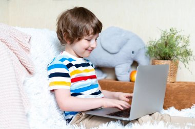 Uzaktan öğrenme çevrimiçi eğitim. Kafkasyalı gülümseyen çocuk evde tablet dizüstü bilgisayarla ders çalışıyor ve okul ödevi yapıyor. Elma ve bilgisayarla kanepede ve battaniyede oturmak.