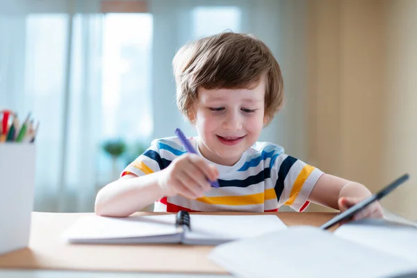 Enseignement à distance en ligne. Blanc sourire garçon enfant étudiant à la maison avec smartphone et faire des devoirs scolaires. Pensant enfant assis à table avec téléphone cellulaire en raison de la quarantaine de coronavirus . — Photo