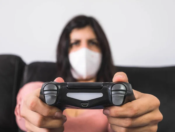 Maske takan bir kadın elinde uzaktan kumandayla kanepede oyun oynuyor.