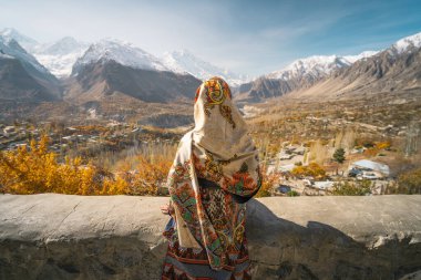 A woman wearing traditional dress sitting on wall and looking at Hunza valley in autumn season, Gilgit Baltistan in Pakistan, Asia clipart