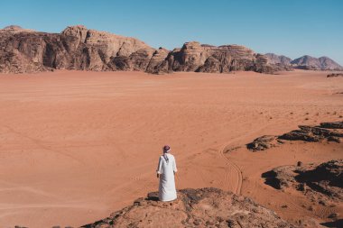 Yerel Arap elbiseli bir adam kayanın üzerinde duruyor ve Ürdün, Arap, Asya 'da Wadi Rum çölüne bakıyor.