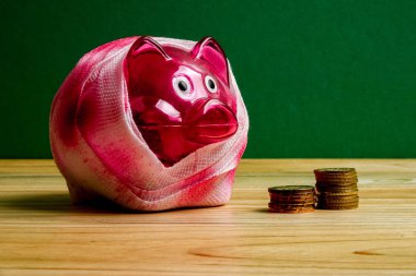 SAVING  DETERIORATE CONCEPT. Red piggy bank with bandage and small stack of coins on the wooden table over green background. clipart