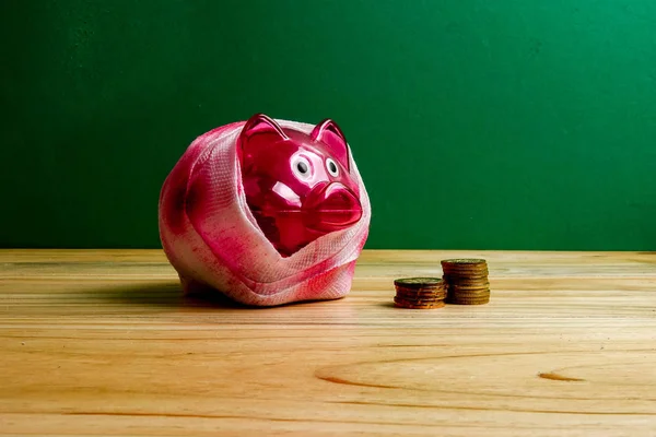 SAVING  DETERIORATE CONCEPT. Red piggy bank with bandage and small stack of coins on the wooden table over green background.