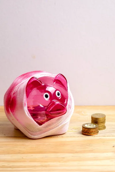 SAVING  DETERIORATE CONCEPT. Red piggy bank with bandage and small stack of coins on the wooden table over white background.