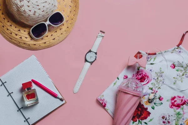 Üst görünüm güneş gözlükleri, şapka, pembe bardak, cam, tişört, kol saati, parfüm, kalem, yaz tatili ve tatil konsepti için pembe arka planda bir defter.