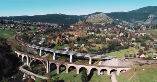 空中風景カラフルな山々とヴォロフタ村 カルパティア山脈 ウクライナの鉄道のヴィアドュートの下に牧歌的な農村の町の上から撮影しました 雄大な風景 — ストック動画
