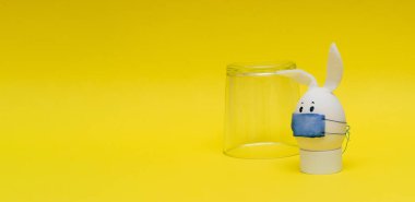 Cam deney şişesi ve gözleri, tavşan kulakları ve sarı arka planda koruyucu bir maskesi olan Paskalya yumurtası. Coronavirüs salgını konsepti. Paskalyayı kendi kendini izole ederek ve karantinaya alarak kutlamayı kabul et
