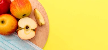 Elma ve koruyucu maskenin tamamı sarı bir arka planda yuvarlak bir kesim tahtasının üzerinde bulunuyor. Coronavirus kavramını hasat edin. Coperid-19 nedeniyle yiyecek kıtlığı kavramı. Metin için boşluk kopyala