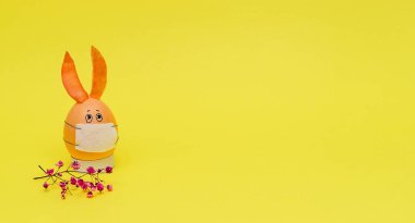 Koruyucu maskeli bir yumurtadan çıkan turuncu Paskalya Tavşanı ve sarı arka planda pembe çingene filizi. Coronavirüs salgını konsepti. Mutlu Paskalya kavramları. Metin için alanı kopyala