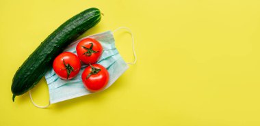 Üç domates ve salatalık sarı bir zemin üzerinde koruyucu bir tıbbi maskenin üzerinde yatıyor. Koronavirüs pandemiyle bağlantılı bir tarım krizi. Koronavirüs konseptini hasat edin.