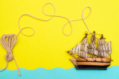 Oyuncak gemi mavi kağıt kesikli dalgalarda yüzüyor, halat sarı arka plandaki bir geminin üstündeki halkalar tarafından bükülüyor. Yazı kavrayın ve düz seyahat edin, taklit alanı oluşturun