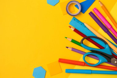 Okul konseptine geri dönelim. Renkli kalemler, keçeli kalemler, cetvel, makas, yapışkan bant ve plastik geometrik şekiller sarı zeminde. Yazı için alanı kopyala, üst görünüm için