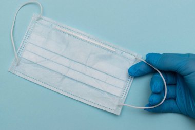 Mavi tıbbi eldivenlerdeki eller mavi arka planda koruyucu bir tıbbi maske tutuyor. Metin için boşluk kopyala, üst görünüm