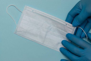 Mavi tıbbi eldivenlerdeki eller mavi arka planda koruyucu bir tıbbi maske tutuyor. Metin için boşluk kopyala, üst görünüm
