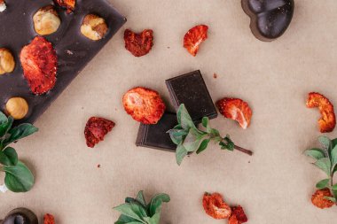  El yapımı koyu çikolatalar ve fındıklı dilimler ve kağıt üzerinde kurutulmuş çilekler. Nane yaprakları ve çilek dilimleri arka plana saçılmış. Sağlıklı tatlılar kavramı, düz uzanma
