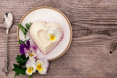 Süzme peynir, agar-agar ve kremalı bir kalp şeklindeki sufle ahşap bir arka planda beyaz bir tabakta. Menekşe çiçekleri kompozisyonu süslüyor. Sağlıklı yiyecek ve sağlıklı tatlı kavramı dışında.