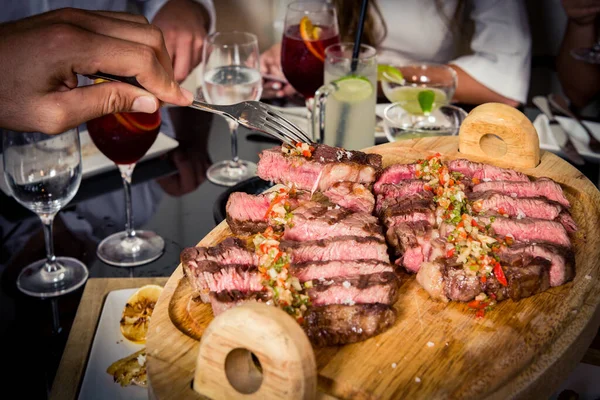 Cut juice steak on a table with friend sharing food, hand with fork, brunch, side view. Beautiful serving of a cut steak with vegetables on a black plate with group of people sharing the food.