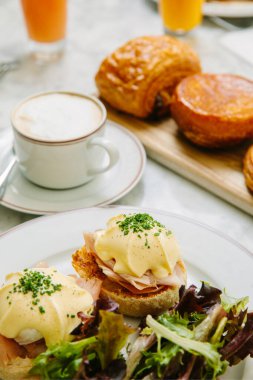 Tabağında salata yaprakları olan Benedict usulü yumurta kahve kapuçino ve hamur işi olan bir restoranda kahvaltı. Kahvaltıda haşlanmış yumurtalı sandviç. Kahve, yemek..