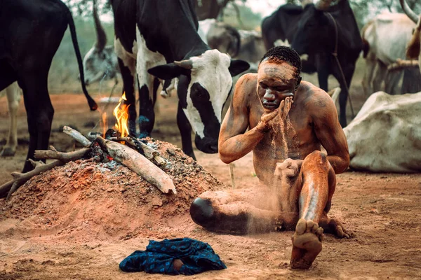 MUNDARI TRIBE, SOUTH SUDAN - 11 marca 2020: Człowiek siedzący na ziemi w pobliżu ogniska i tradycyjnie smarowanie ciała popiołem pomarańczowym dla higieny podczas wypasu zwierząt z innymi plemionami w sawannie — Zdjęcie stockowe