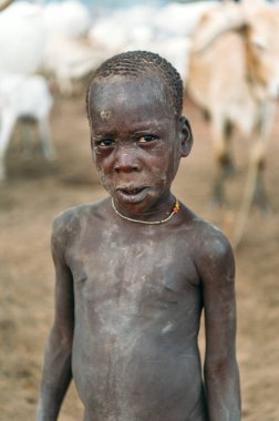 MUNDARI TRIBE, GÜNEY SUDAN - 11 Mart 2020: Güney Sudan 'daki otlakta bulanık inek sürüsünün karşısında duran çıplak gövdeli çocuk kameraya bakıyor