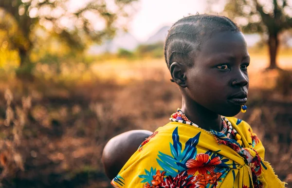 BOYA TRIBE, SOUTH SUDAN - MARCH 10, 2020: Girl in traditional colorful garment of Boya Tribe carrying baby on back on blurred background of savanna in South Sudan, Africa — Stock Photo, Image