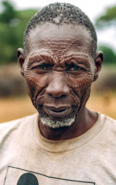 TOPOSA TRIBE, Güney Sudan, Afrika 'da Toposa Kabile Köyü' nde yaşarken yüzünde geleneksel yara izleri olan ve kirli tişörtlü yaşlı bir erkek.