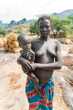 BOYA TRIBE, Güney SUDAN - 10 Mart 2020: Geleneksel renkli giysiler ve aksesuarlı kadın ayakta dururken ve kucağında sakin küçük bir çocuğu taşırken kameraya ilgi ile bakıyor