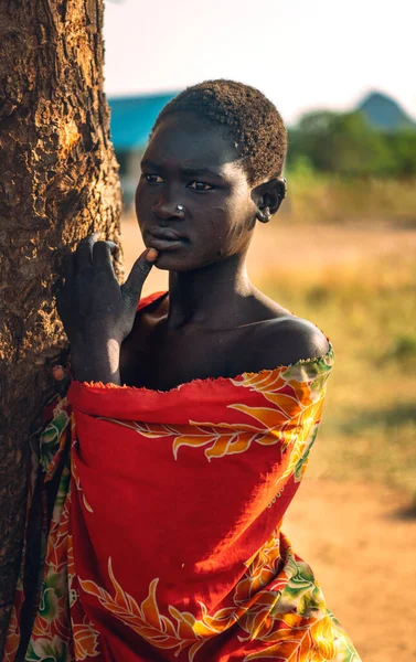 BOYA TRIBE, SOUTH SUDAN - 10 marca 2020: Młoda kobieta w kolorowym ubraniu dotykająca ust i odwracająca wzrok, opierająca się na pniu sawanny w pobliżu wioski Boya Tribe w Sudanie Południowym, Afryka — Zdjęcie stockowe