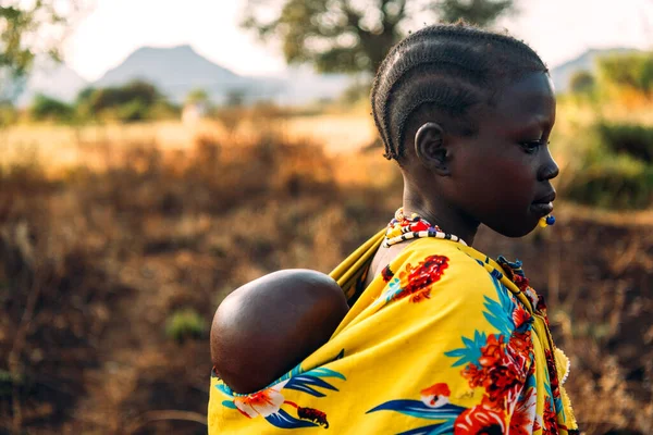 BOYA TRIBE, SOUTH SUDAN - 10 marca 2020: Dziewczyna w tradycyjnej kolorowej odzieży plemienia Boya nosząca dziecko na plecach na rozmytym tle sawanny w Sudanie Południowym, Afryka — Zdjęcie stockowe