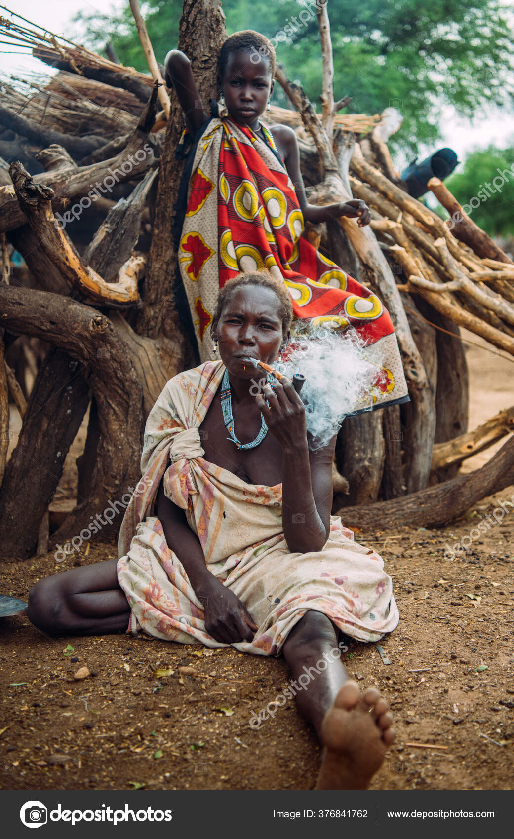 TRIBE, SOUTH SUDAN - MARCH 12, 2020: Kvinde rygerør, mens hun sidder på jorden nær tømmer og pige i farverigt tøj i landsbyen Toposa Tribe i Sydsudan, Afrika Redaktionelle stock-fotos © klublub