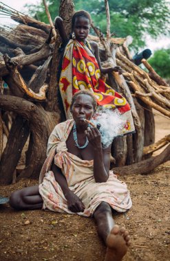 TOPOSA TRIBE, Güney SUDAN - 12 Mart 2020: Afrika 'nın güneyindeki Toposa Kabilesi köyünde kereste ve kız renkli giysiler içinde yerde otururken sigara içen kadın