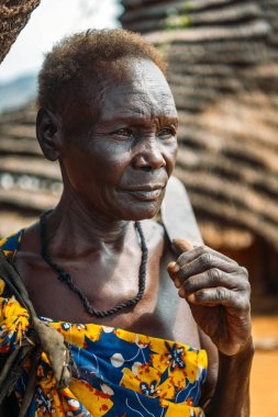 BOYA TRIBE, Güney Sudan - 10 Mart 2020: Kısa saçlı, omuzlarında bıçak taşıyan ve Güney Sudan 'daki Boya Kabilesi köyünün bulanık arka planına bakan yaşlı kadın